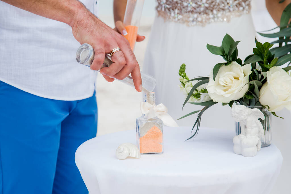Νύφη και γαμπρός ρίχνει πολύχρωμα διαφορετικά χρώματα άμμου στο κρυστάλλινο βάζο κοντά κατά τη διάρκεια συμβολική ναυτική διακόσμηση προορισμός γαμήλια τελετή στην αμμώδη παραλία μπροστά από τον ωκεανό  - Φωτογραφία, εικόνα