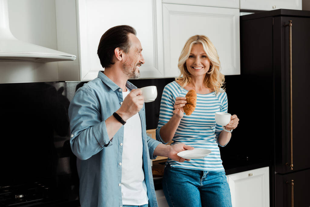 笑顔女性保持クロワッサン近く夫とともにコーヒーカップでキッチン  - 写真・画像