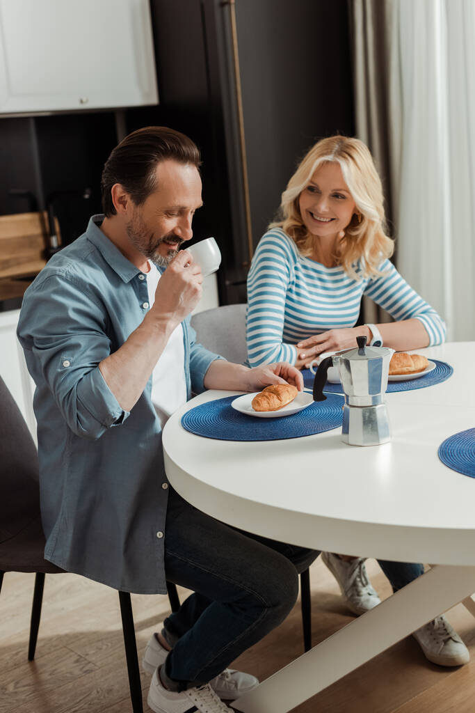 Komea mies juo kahvia lähellä hymyilevä vaimo pöydässä keittiössä  - Valokuva, kuva