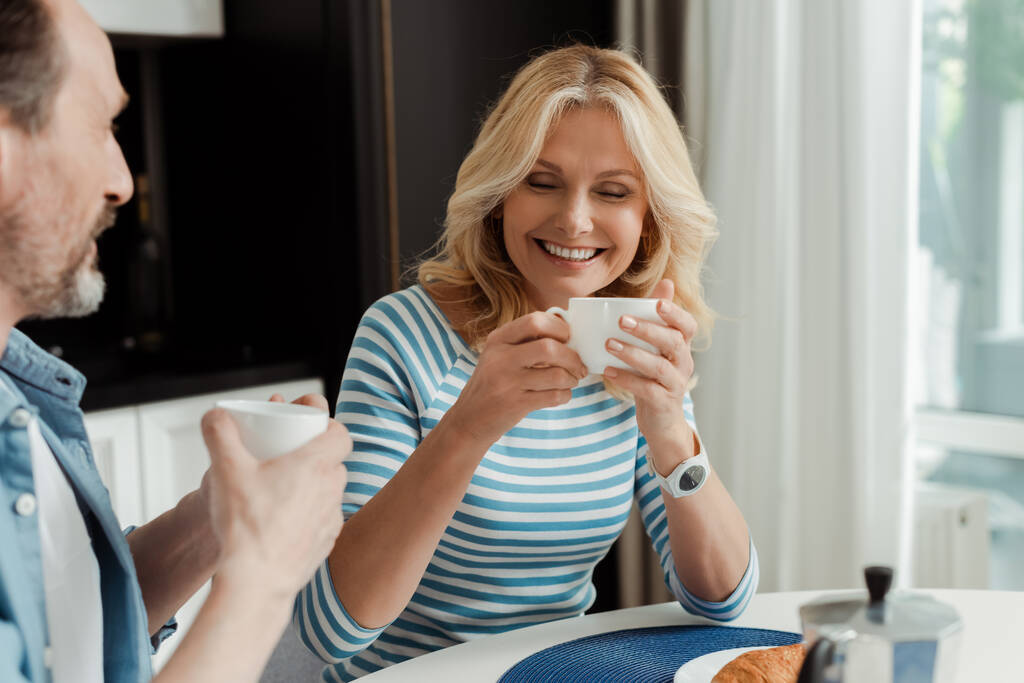 Επιλεκτική εστίαση της όμορφης γυναίκας χαμογελώντας πίνοντας καφέ με τον σύζυγο στο σπίτι  - Φωτογραφία, εικόνα