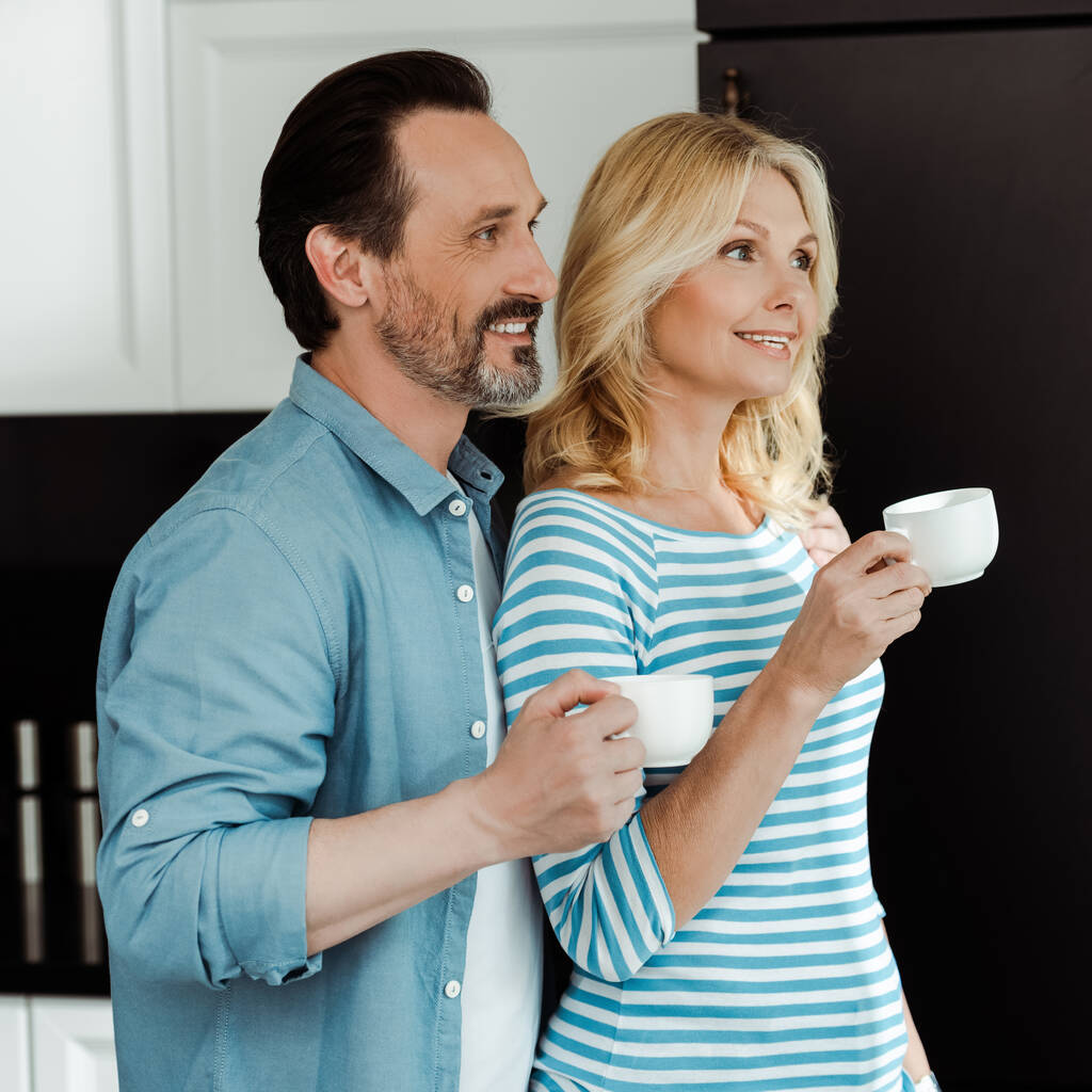 Glimlachende man in de buurt van mooie vrouw met kopje koffie thuis  - Foto, afbeelding