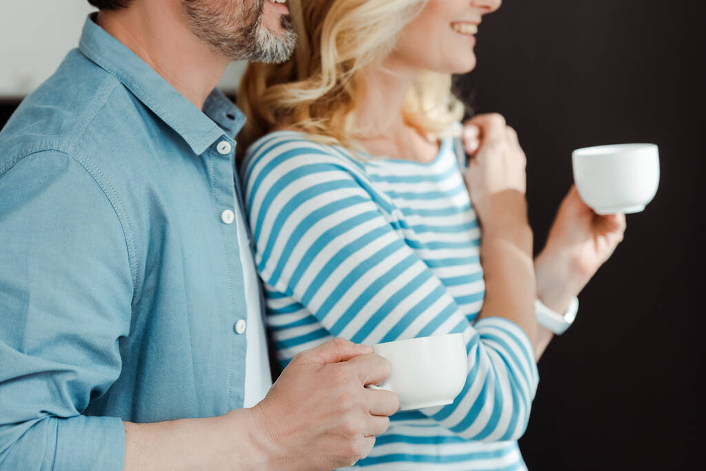Vista recortada del hombre abrazando esposa sonriente con taza de café en casa
  - Foto, Imagen