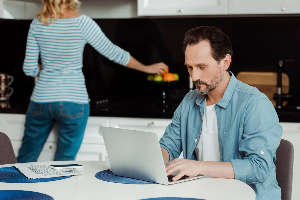 Mann benutzt Laptop, während Frau Orange in Küche nimmt  - Foto, Bild