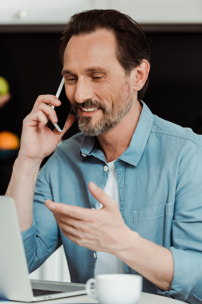 Вибірковий фокус усміхненого чоловіка, який розмовляє на мобільному телефоні біля ноутбука та кави на кухонному столі
  - Фото, зображення