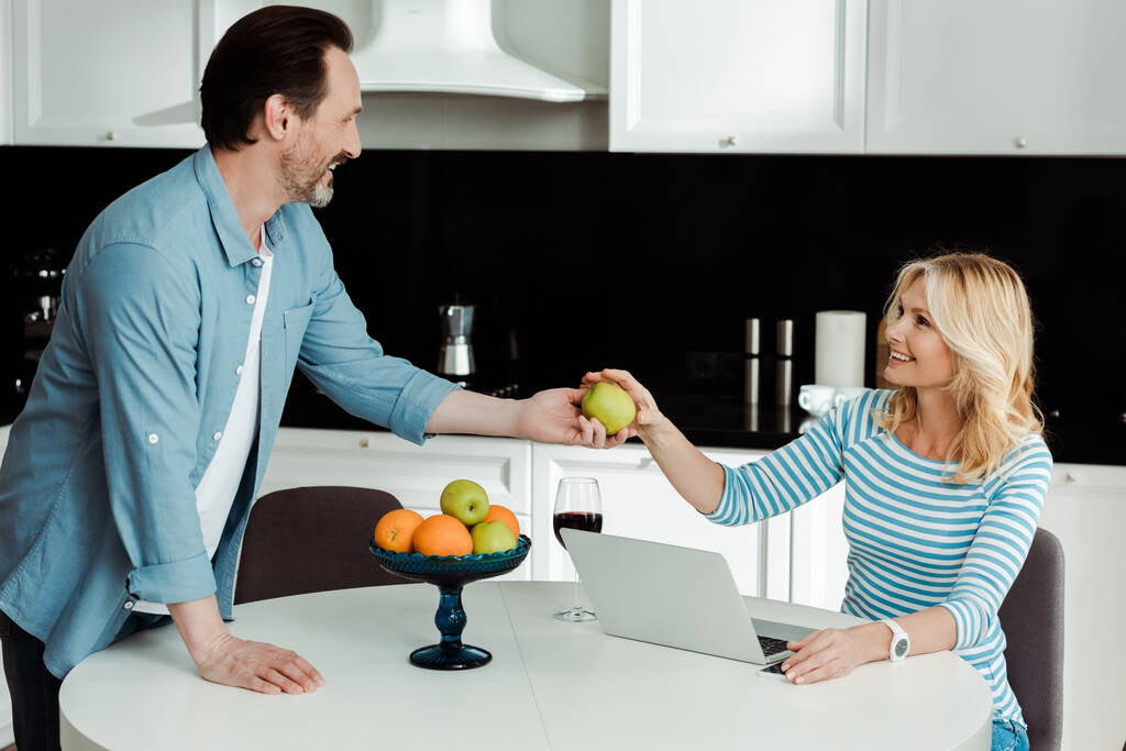 Sonriente hombre dando manzana a la esposa cerca de vino y portátil en la mesa de la cocina
  - Foto, imagen