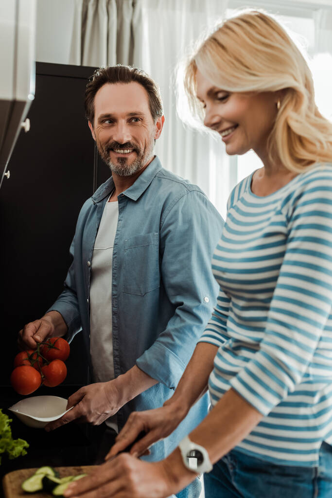 Выборочный фокус улыбающегося мужчины, смотрящего на жену во время приготовления салата на кухне
  - Фото, изображение