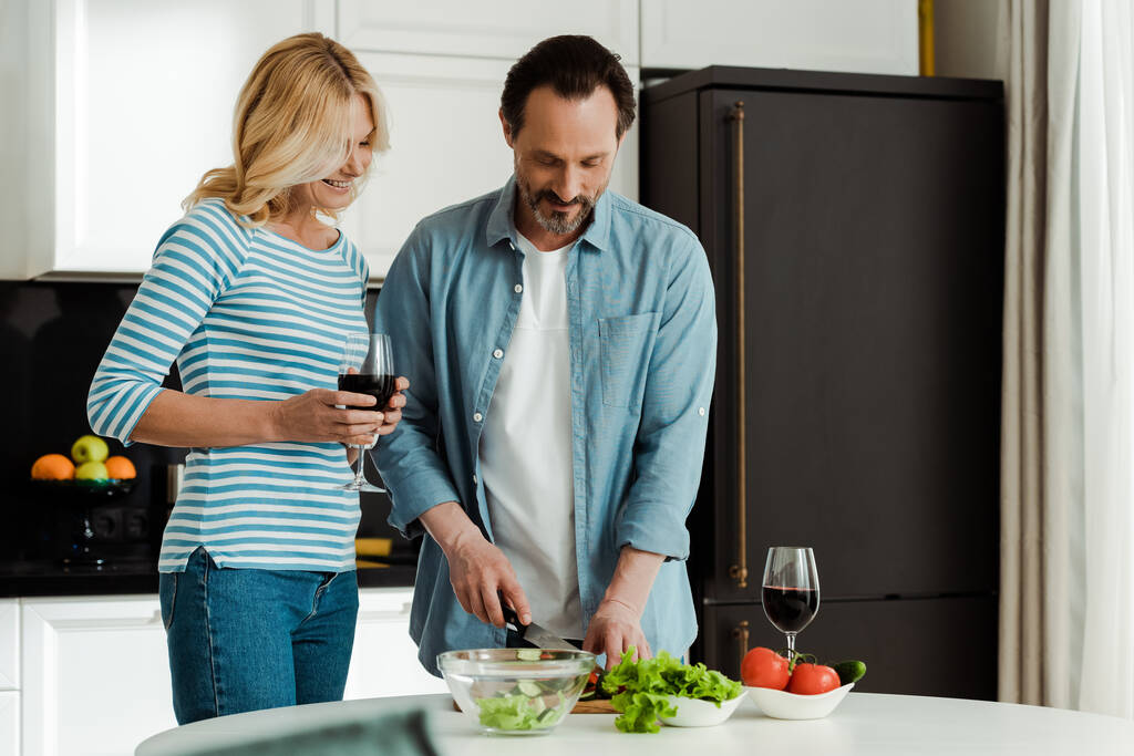 Выборочный фокус улыбающейся женщины, держащей бокал вина рядом с мужем, готовящим салат на кухне
  - Фото, изображение