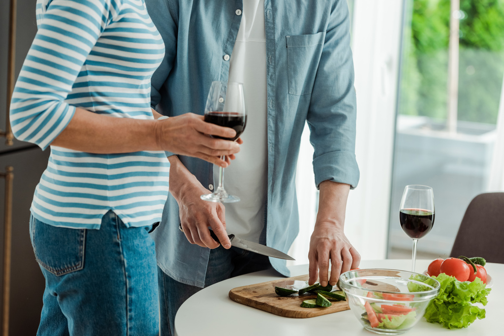 Обрізаний вид жінки, що тримає келих вина біля чоловіка, що ріже овочі на кухні
  - Фото, зображення