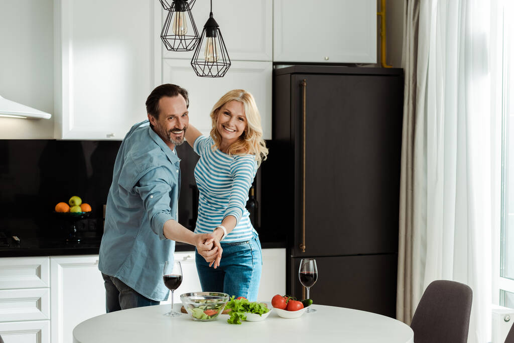 Vrolijk volwassen paar dansen in de buurt van wijnglazen en verse salade in de keuken  - Foto, afbeelding