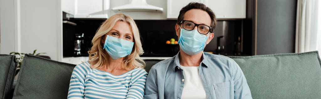 Panoramic shot of mature couple in medical masks sitting on couch at home  - Photo, Image