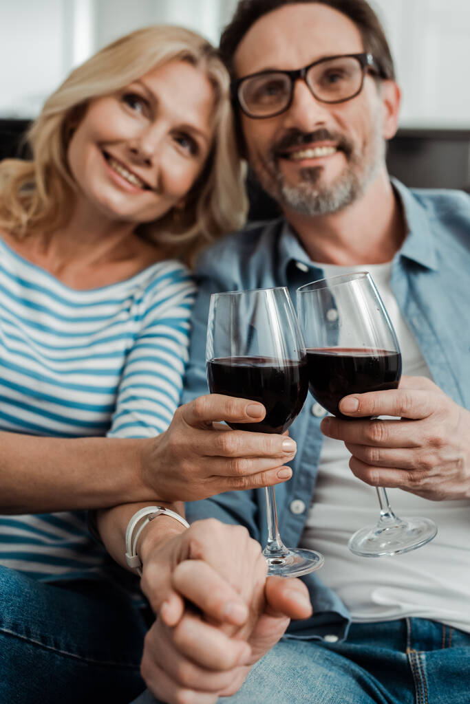 Selektywne skupienie się uśmiechniętej dojrzałej pary trzymającej się za ręce i brzęczącej winem w domu  - Zdjęcie, obraz