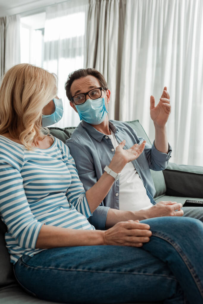 Selektiver Fokus der Frau in medizinischer Maske, die mit der Hand in der Nähe ihres Mannes auf der Couch zeigt  - Foto, Bild
