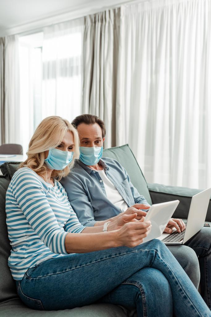 Middelbaar koppel in medische maskers met behulp van digitale tablet en laptop in de woonkamer - Foto, afbeelding