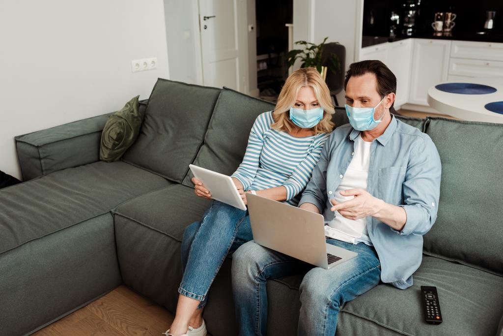 Mężczyzna w masce medycznej wskazując na laptopa w pobliżu żony z tabletem cyfrowym na kanapie  - Zdjęcie, obraz