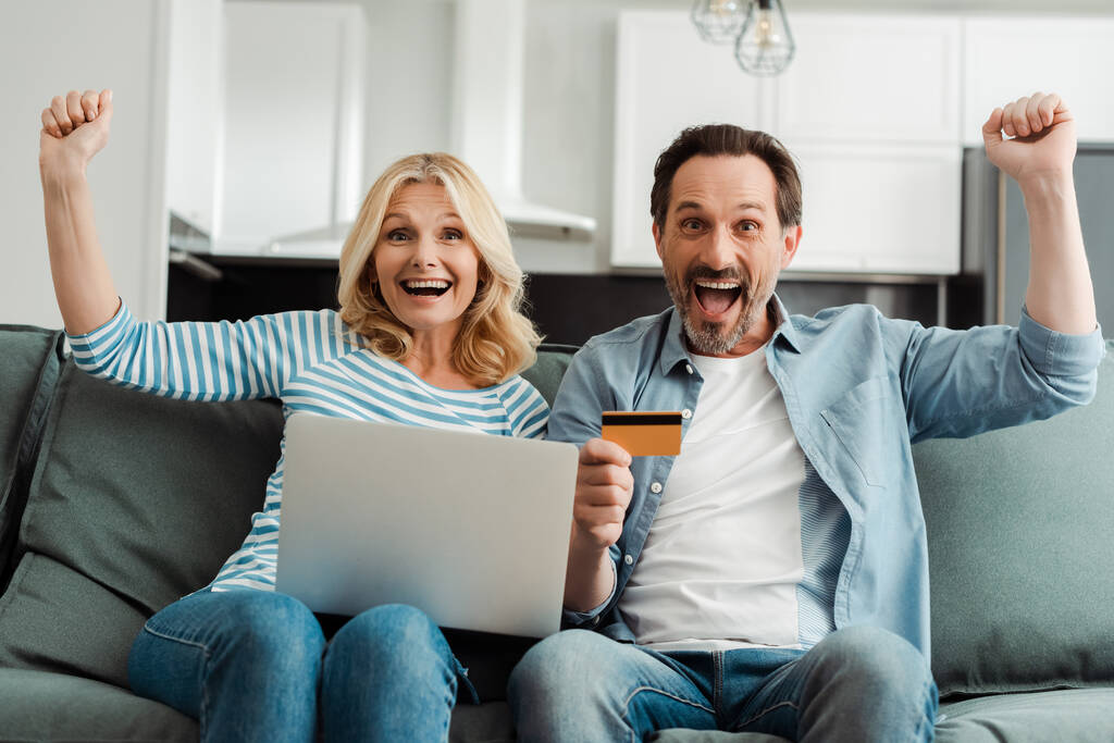 Emocionada pareja madura mostrando sí gesto mientras se utiliza la tarjeta de crédito y el ordenador portátil en el sofá
  - Foto, imagen
