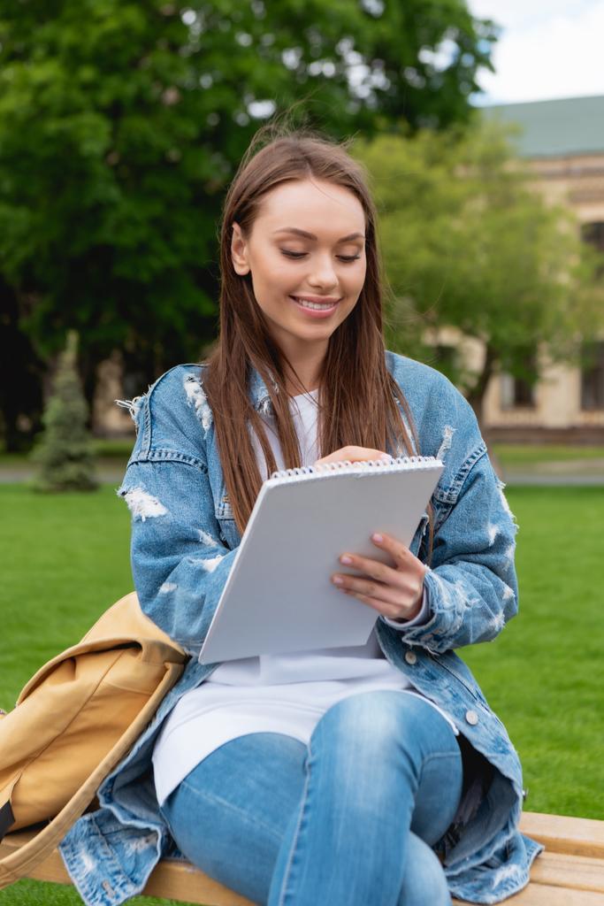 fröhliche junge Studentin schreibt in Notizbuch im Park  - Foto, Bild