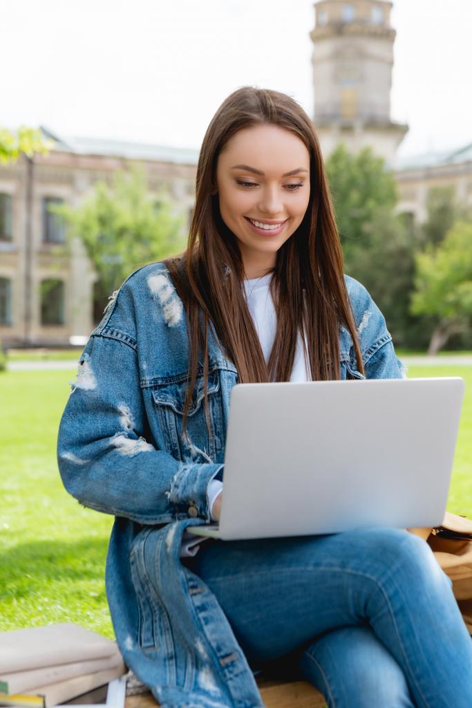 χαρούμενα φοιτητής κάθεται στον πάγκο και τη χρήση φορητού υπολογιστή κοντά σε βιβλία, online έννοια μελέτη  - Φωτογραφία, εικόνα