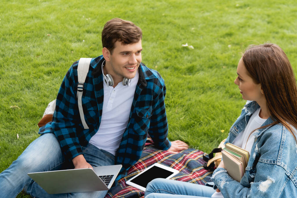 glücklicher Student schaut attraktives Mädchen in der Nähe von Gadgets auf karierter Decke an  - Foto, Bild
