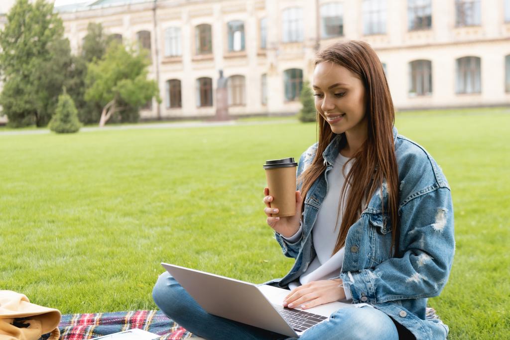 ευτυχής και ελκυστικός μαθητής που κρατά το κύπελλο μιας χρήσης ενώ χρησιμοποιεί το lap-top, σε απευθείας σύνδεση έννοια μελέτη  - Φωτογραφία, εικόνα