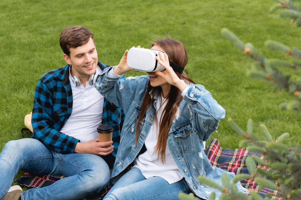 selektywne skupienie radosnego studenta trzymającego papierową filiżankę i patrzącego na podekscytowaną dziewczynę w wirtualnej rzeczywistości gogli  - Zdjęcie, obraz