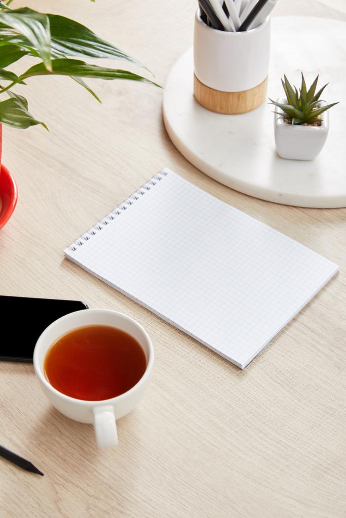 grüne Pflanzen, Tasse Tee und leeres Notizbuch in der Nähe des Smartphones auf hölzerner Oberfläche - Foto, Bild