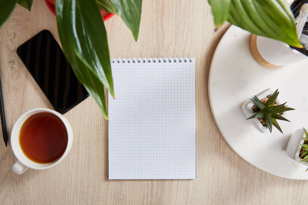 Blick von oben auf grüne Pflanzen, Tasse Tee und leeres Notizbuch in der Nähe von Smartphone auf Holzoberfläche - Foto, Bild