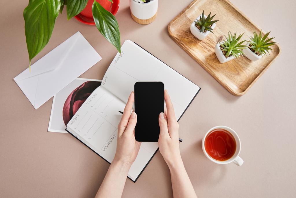 widok z góry żeńskich rąk ze smartfonem w pobliżu zielonych roślin na drewnianej desce, filiżanka herbaty, planista z ołówkiem na beżowej powierzchni - Zdjęcie, obraz