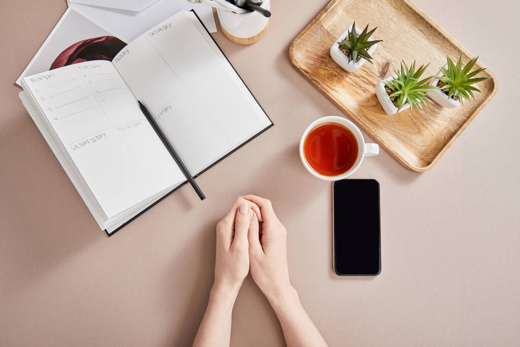bovenaanzicht vrouwelijke handen, smartphone bij groene planten op houten bord, kopje thee, planner met potlood op beige oppervlak - Foto, afbeelding
