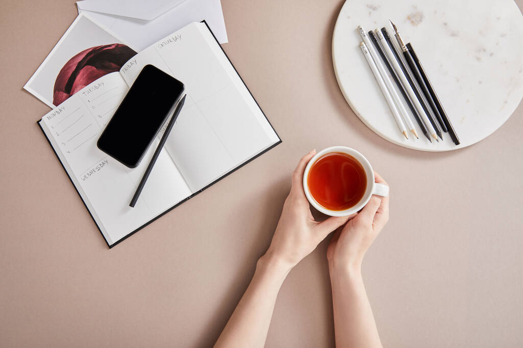 Planlayıcı, kart, akıllı telefon yakınında elinde çay fincanı tutan bir kadının bej rengi yüzeyi kalemle daire içine alınmış görüntüsü. - Fotoğraf, Görsel