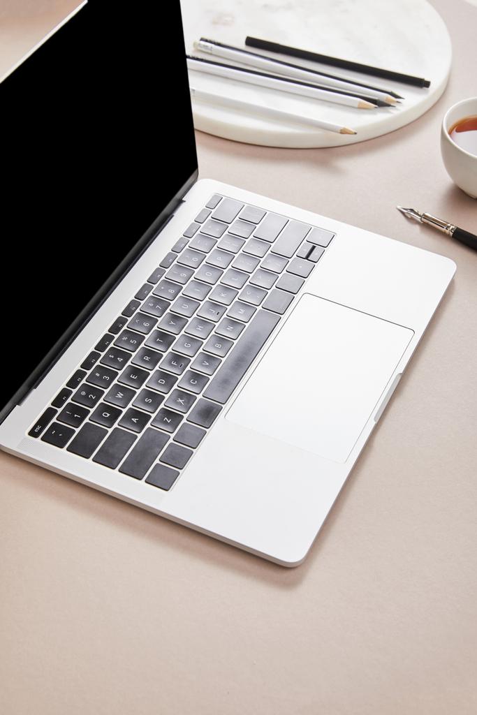 laptop με κενή οθόνη κοντά στο φλιτζάνι τσάι και χαρτικά σε μπεζ επιφάνεια - Φωτογραφία, εικόνα