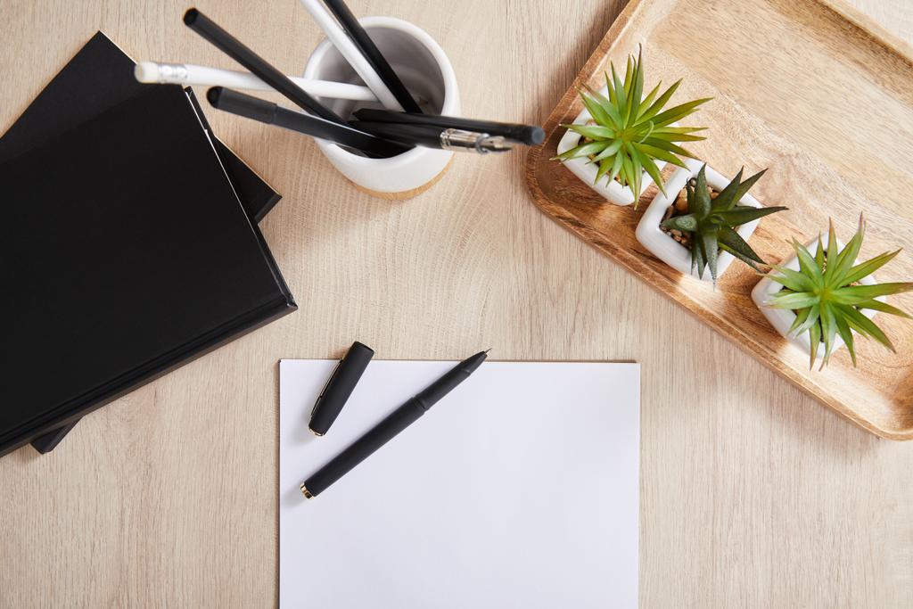 shora pohled na zelené rostliny, černé zápisníky, tužky v držáku a bílý papír s perem na dřevěném povrchu - Fotografie, Obrázek