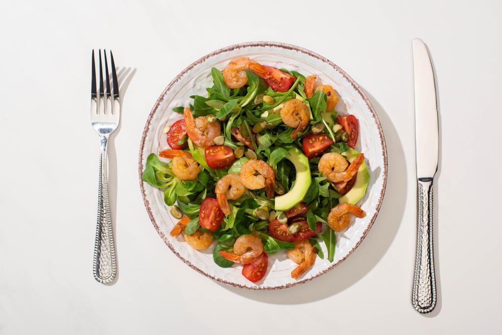top view of fresh green salad with shrimps and avocado on plate near cutlery on white background - Photo, Image