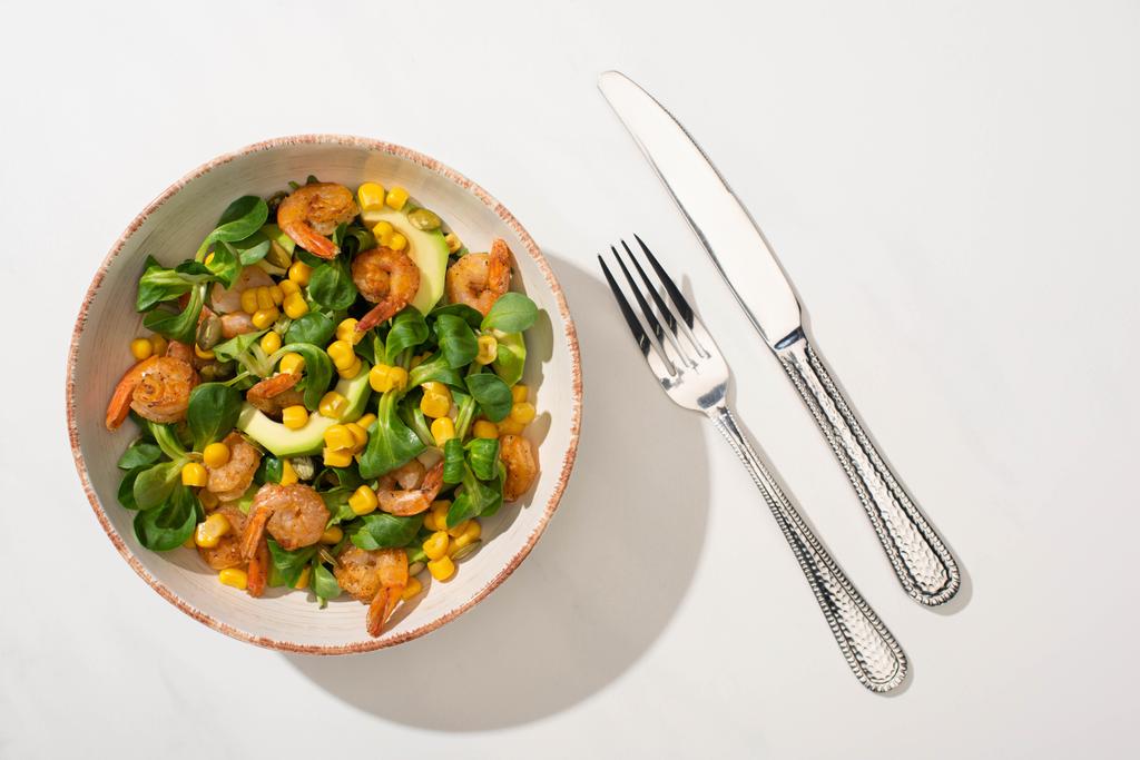Draufsicht auf frischen grünen Salat mit Mais, Garnelen und Avocado auf Teller neben Besteck auf weißem Hintergrund - Foto, Bild