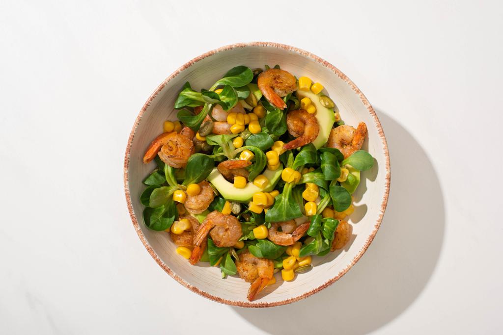 Draufsicht auf frischen grünen Salat mit Mais, Garnelen und Avocado auf Teller auf weißem Hintergrund - Foto, Bild
