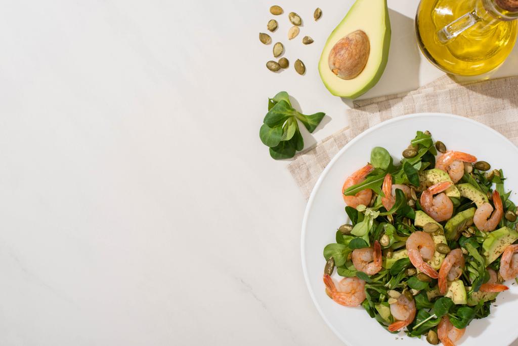вид на свежий зеленый салат с тыквенными семечками, креветками и авокадо на тарелке на салфетке рядом с ингредиентами на белом фоне
 - Фото, изображение