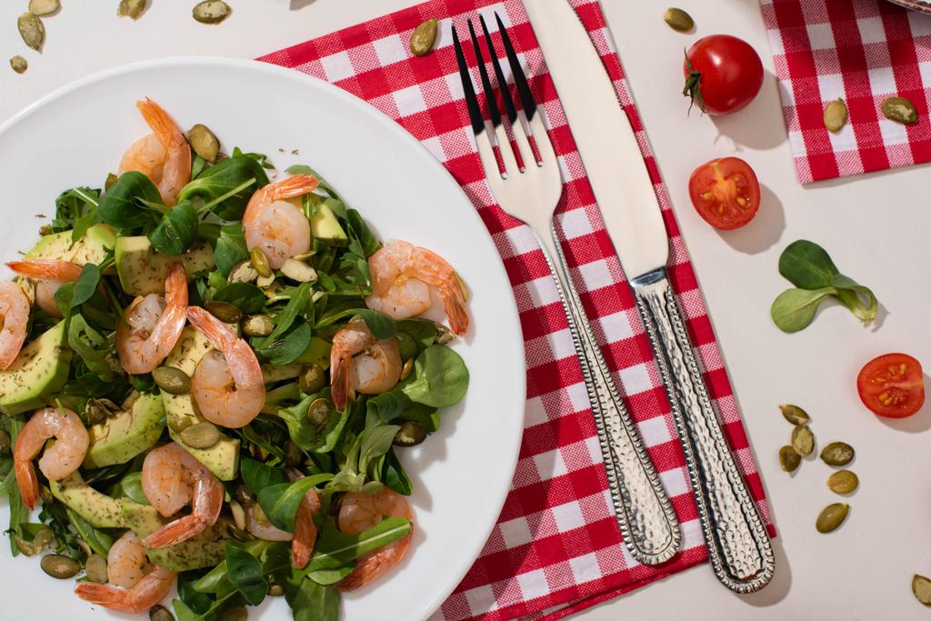 top view of fresh green salad with shrimps and avocado on plate near cutlery on plaid napkin and ingredients on white background - Photo, Image