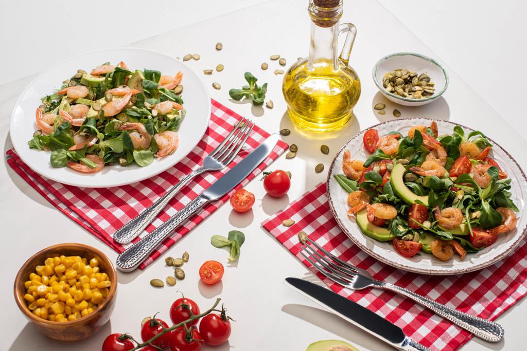 salada verde fresca com camarões e abacate em pratos perto de talheres em guardanapos xadrez e ingredientes na mesa branca
 - Foto, Imagem
