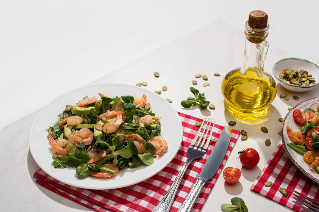 fresh green salad with shrimps and avocado on plate near cutlery on plaid napkin and ingredients on white table - Photo, Image