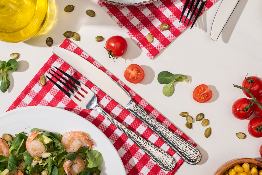 Draufsicht auf frischen grünen Salat mit Garnelen und Avocado auf Teller in der Nähe von Besteck auf karierter Serviette und Zutaten auf weißem Hintergrund - Foto, Bild
