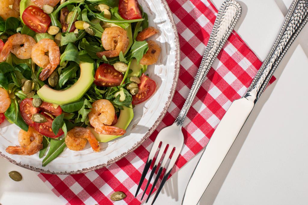 vista dall'alto di insalata verde fresca con gamberetti e avocado su piatto vicino a posate su tovagliolo a quadri su sfondo bianco
 - Foto, immagini