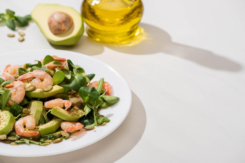 selektivní zaměření čerstvého zeleného salátu s krevetami a avokádem na talíři v blízkosti olivového oleje na bílém pozadí - Fotografie, Obrázek