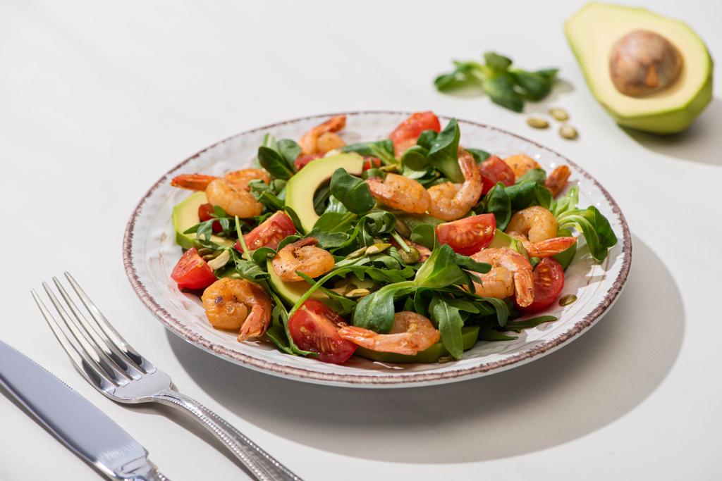 selektiver Schwerpunkt von frischem grünem Salat mit Garnelen und Avocado auf Teller neben Besteck auf weißem Hintergrund - Foto, Bild