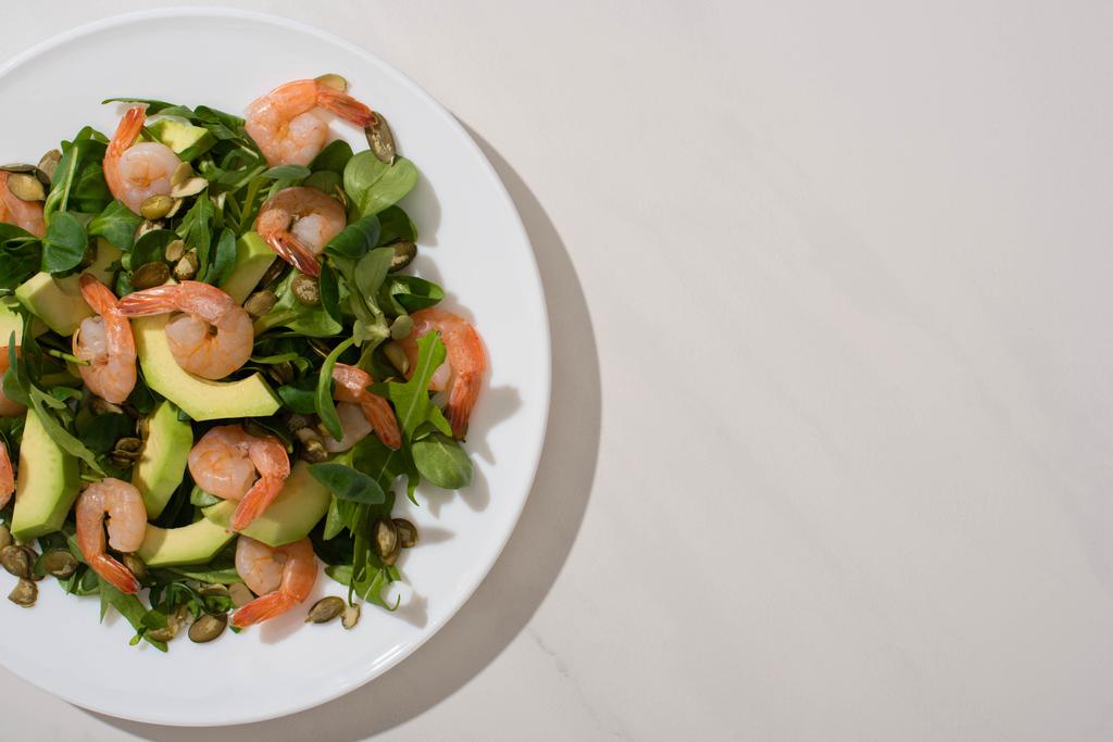 вид зверху свіжий зелений салат з гарбузовим насінням, креветками та авокадо на тарілці на білому фоні
 - Фото, зображення