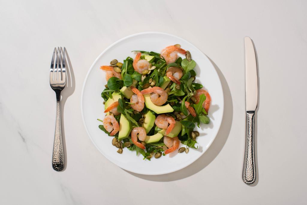 πάνω άποψη του φρέσκια πράσινη σαλάτα με σπόρους κολοκύθας, γαρίδες και αβοκάντο στο πιάτο κοντά μαχαιροπήρουνα σε λευκό φόντο - Φωτογραφία, εικόνα