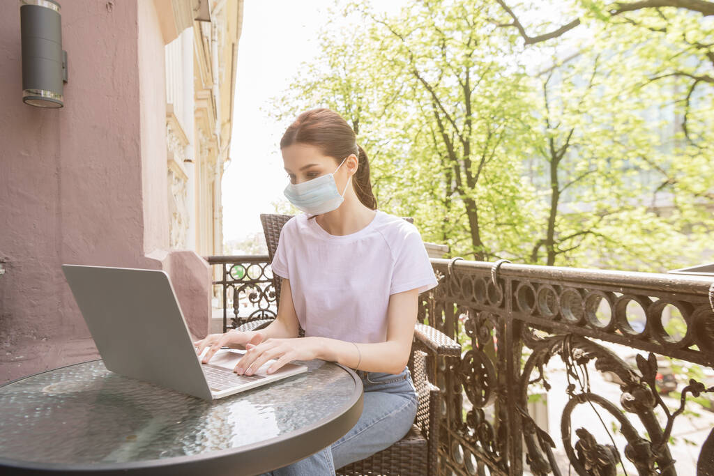 Junge Freiberuflerin in medizinischer Maske sitzt auf Stuhl und benutzt Laptop auf Balkon  - Foto, Bild