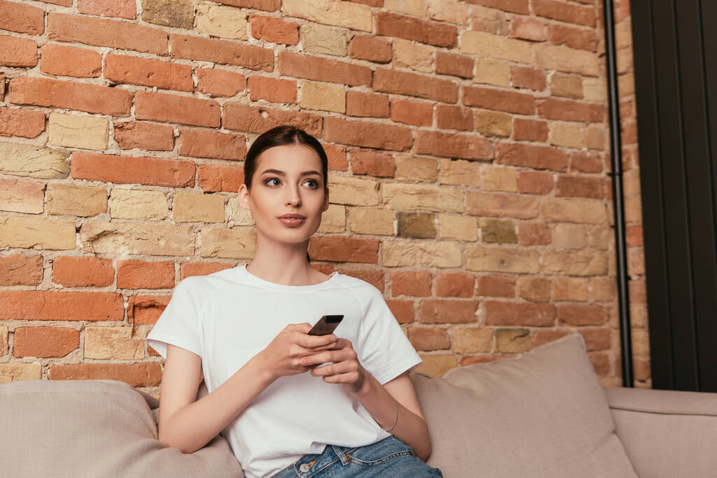attractive young woman sitting on sofa and holding remote controller  - Photo, Image