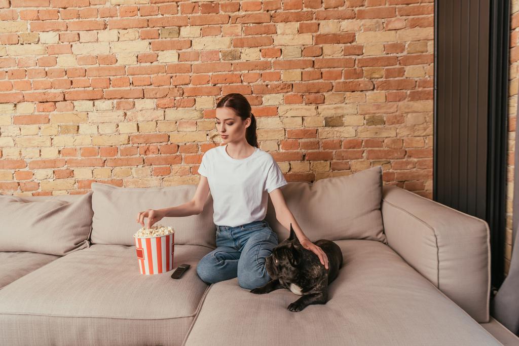 donna felice raggiungendo popcorn e seduto sul divano con carino bulldog francese
 - Foto, immagini