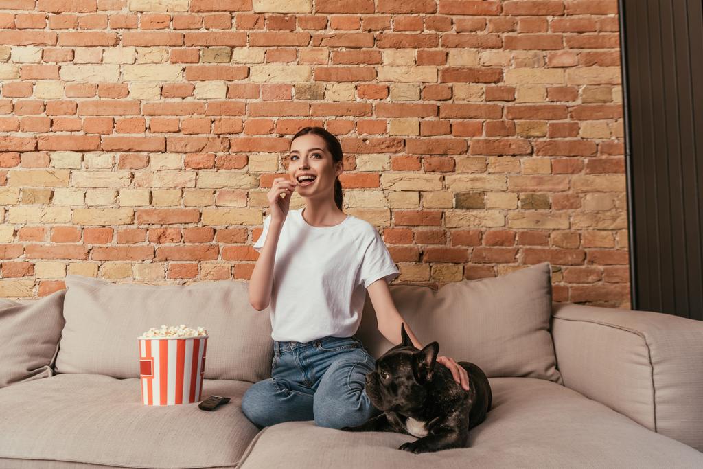 happy woman eating popcorn and sitting on sofa with cute french bulldog while watching movie - Photo, Image