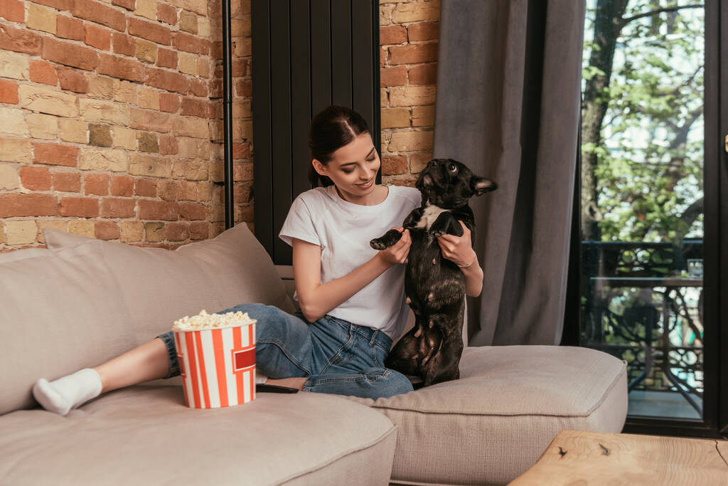 boldog lány ül a kanapén közel popcorn vödör és megható aranyos francia bulldog  - Fotó, kép