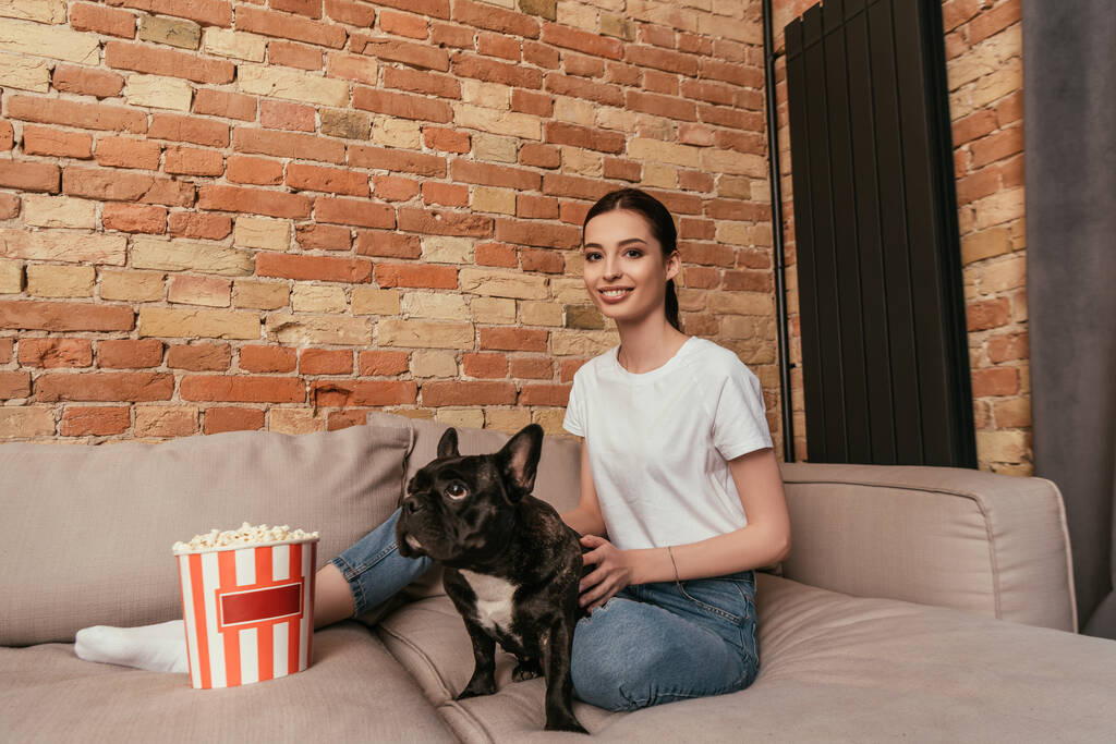 hymyilevä nainen istuu sohvalla lähellä popcorn ämpäri ja koskettaa söpö ranskalainen bulldog  - Valokuva, kuva